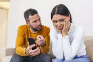 Duas pessoas estão sentadas no sofá. Uma delas aponta o celular e olha para a outra, que tampa as orelhas com as mãos e está emburrada.