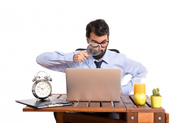 Um homem usando camisa social e gravata está apontando uma lupa para o computador em sua mesa.