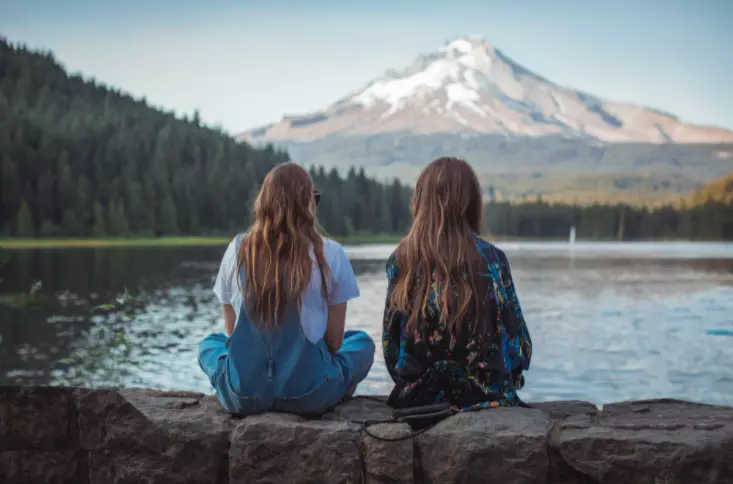 duas mulheres sentadas em um muro de pedra olhando para um lago