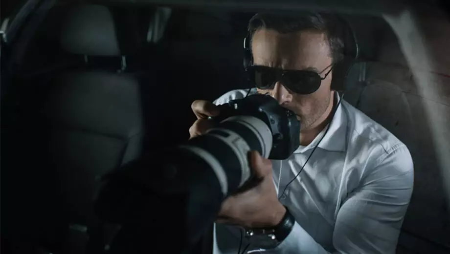 homem detetive particular, de óculos escuro, dentro de um carro, com fones de ouvido e uma câmera profissional de fotografia e filmagem fazendo um trabalho investigativo